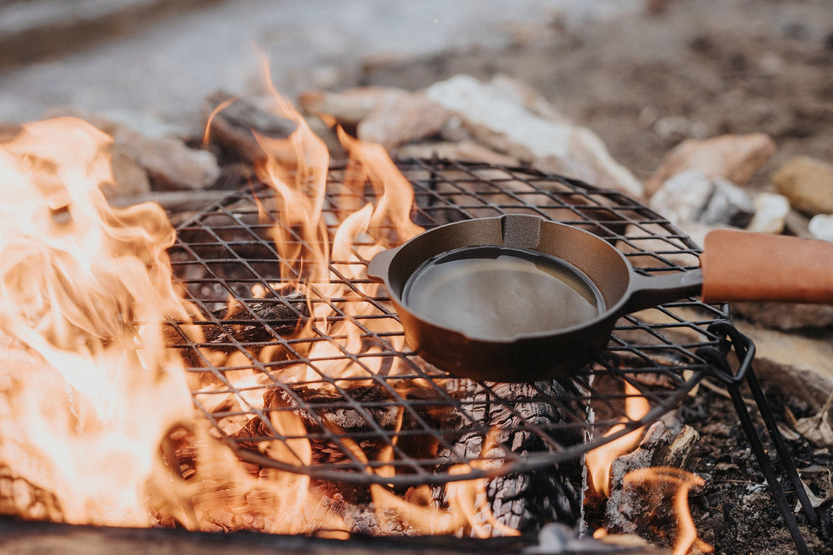 Small cast iron skillet over a small circular grill grate on a campfire with flames and rocks in the background