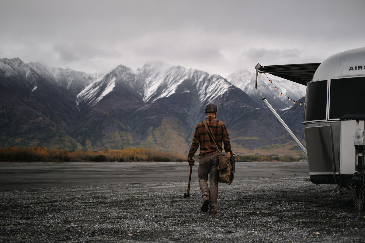 Man walking towards a camper with mountains in the background carrying Barebones products